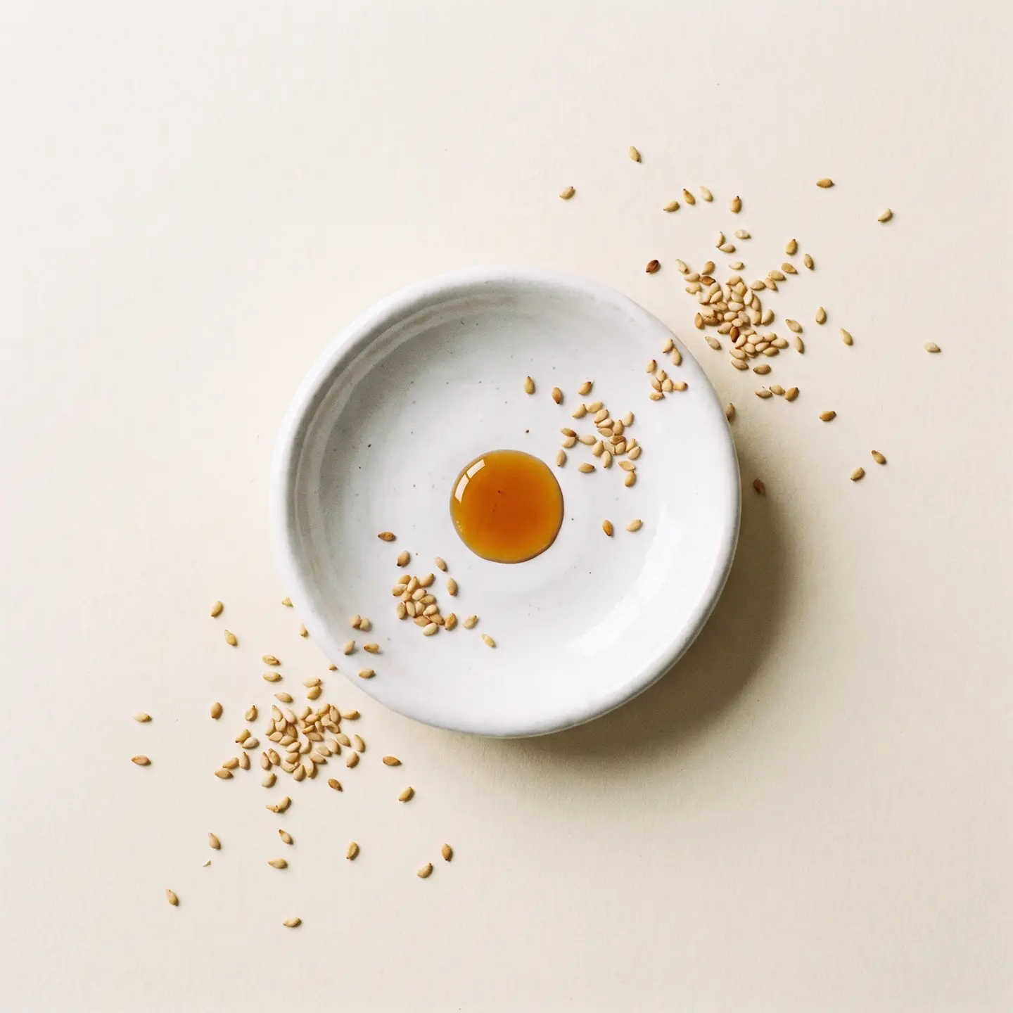 A single amber drop of oregano oil falling into a white ceramic dish ringed with sesame seeds