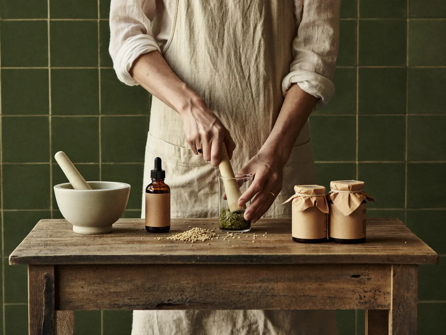 Hands cold-pressing oregano at a wooden atelier bench, sesame seeds and amber glass nearby